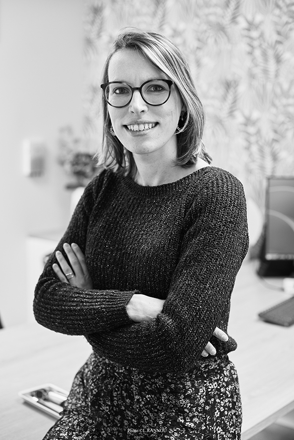 Portrait en noir et blanc d'Audrey, gérante souriante de Audition Conseil à Plescop, portant des lunettes et posant au centre des 3 Soleils.